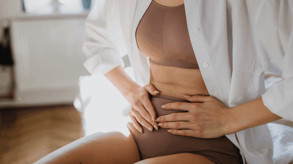 A woman sitting on a bed holding her lower abdomen, representing the pain and discomfort some women experience after childbirth.