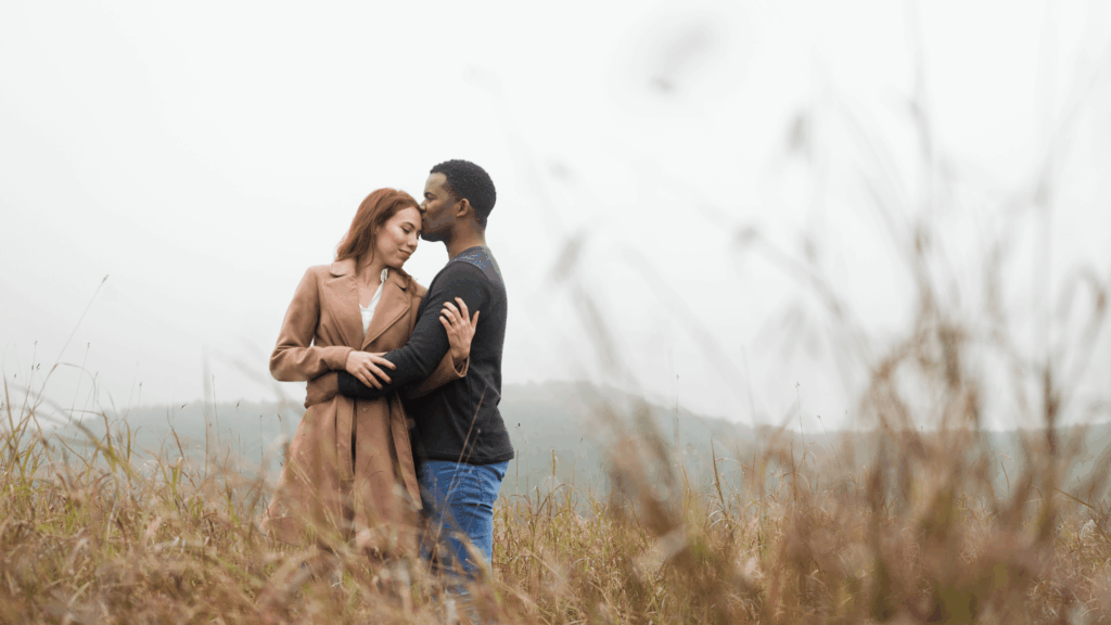 A couple stands embracing in a field, with the man kissing the woman's forehead, symbolizing a restored sense of intimacy and connection.